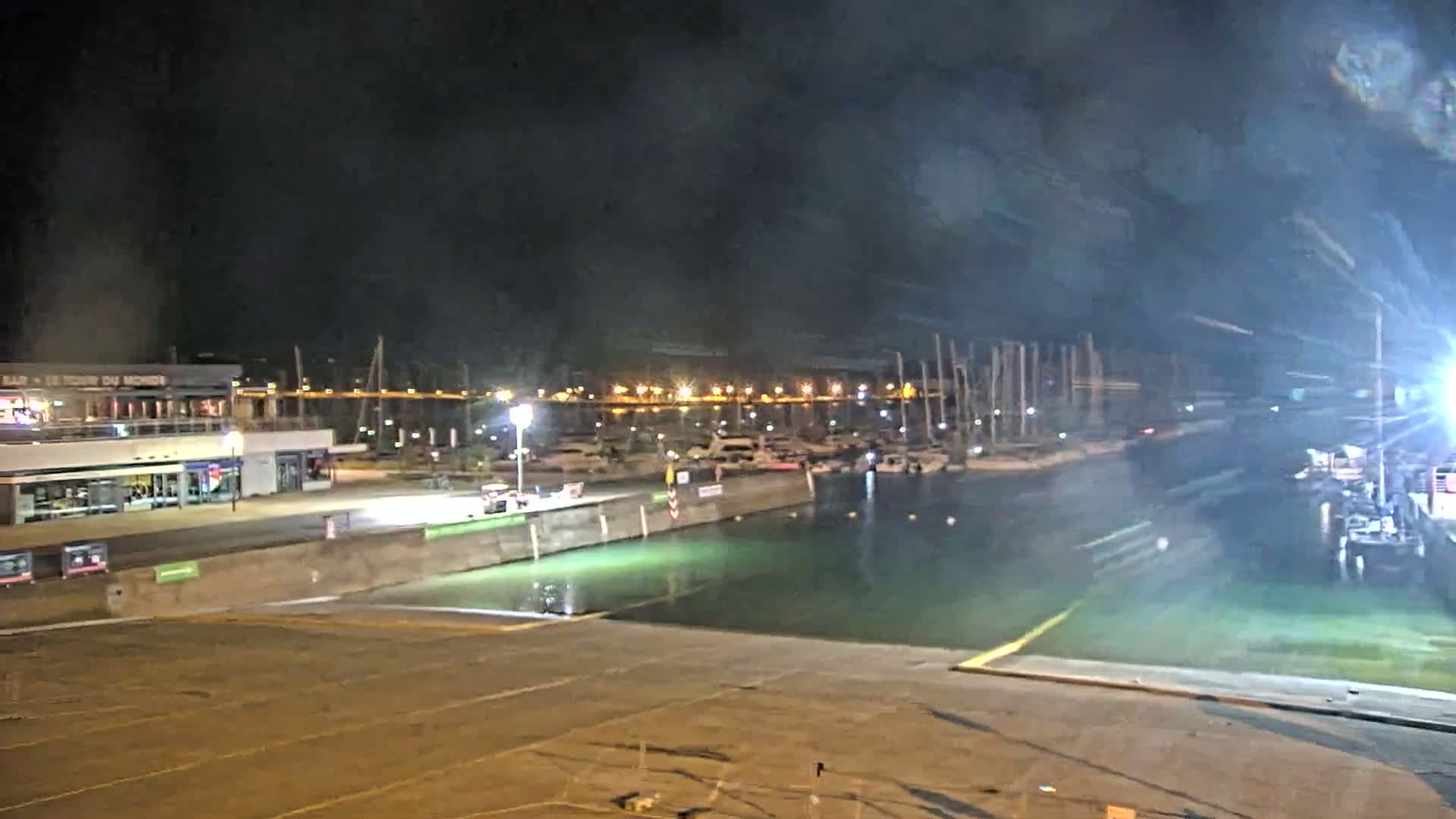 A nighttime view of a marina with docked boats, illuminated buildings, and a calm, greenish body of water.