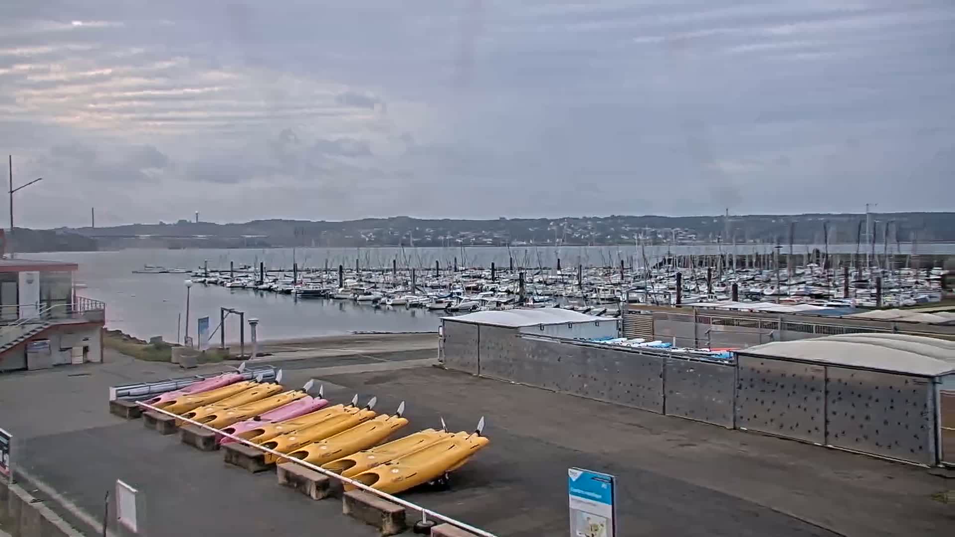 A marina filled with sailboats under a cloudy sky, with several kayaks lined up on the shore.