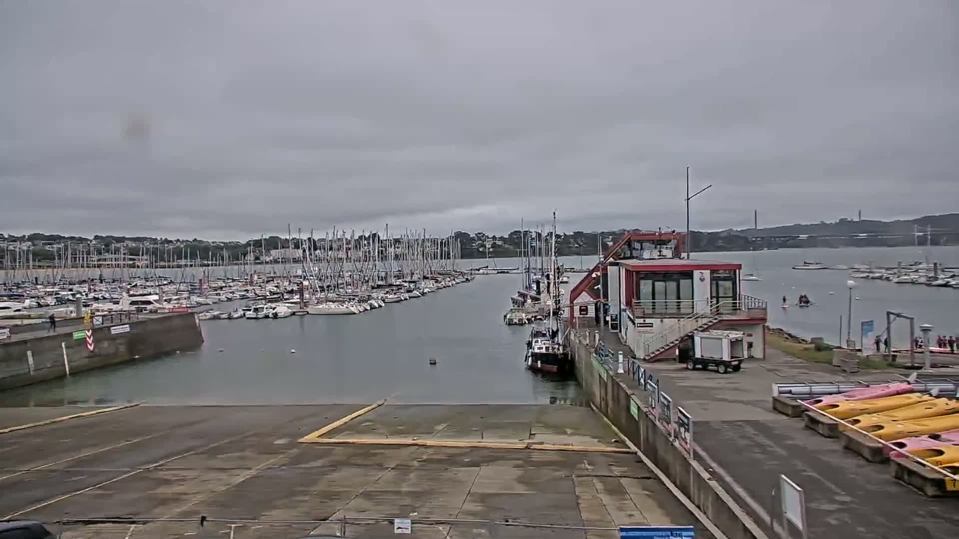 A marina filled with sailboats under a cloudy sky, with a boat ramp in the foreground and kayaks lined up on the shore.