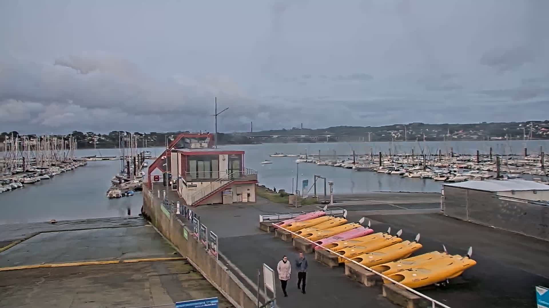 Two people walk past rows of yellow and pink kayaks near a marina filled with boats under an overcast sky.