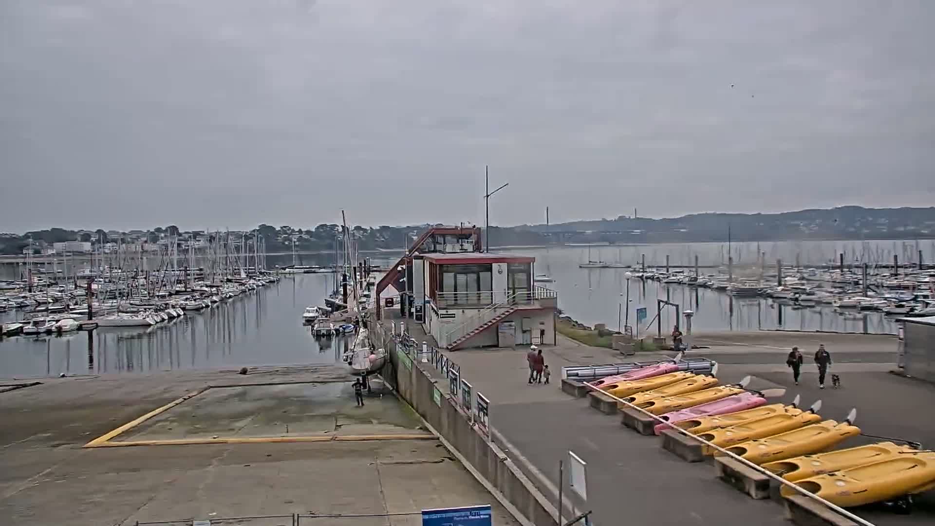 A marina filled with sailboats under an overcast sky, with a small building, several kayakers, and a few people walking nearby.