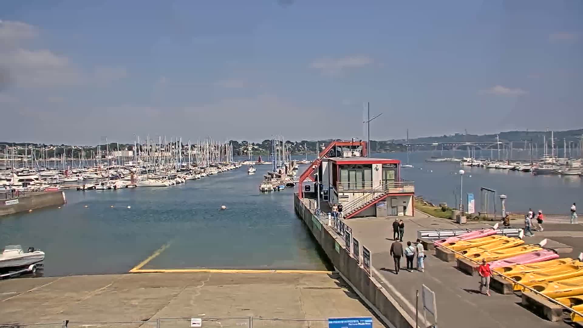 A marina filled with sailboats and motorboats on a sunny day, with a modern red building and rows of kayaks situated on a paved area nearby.