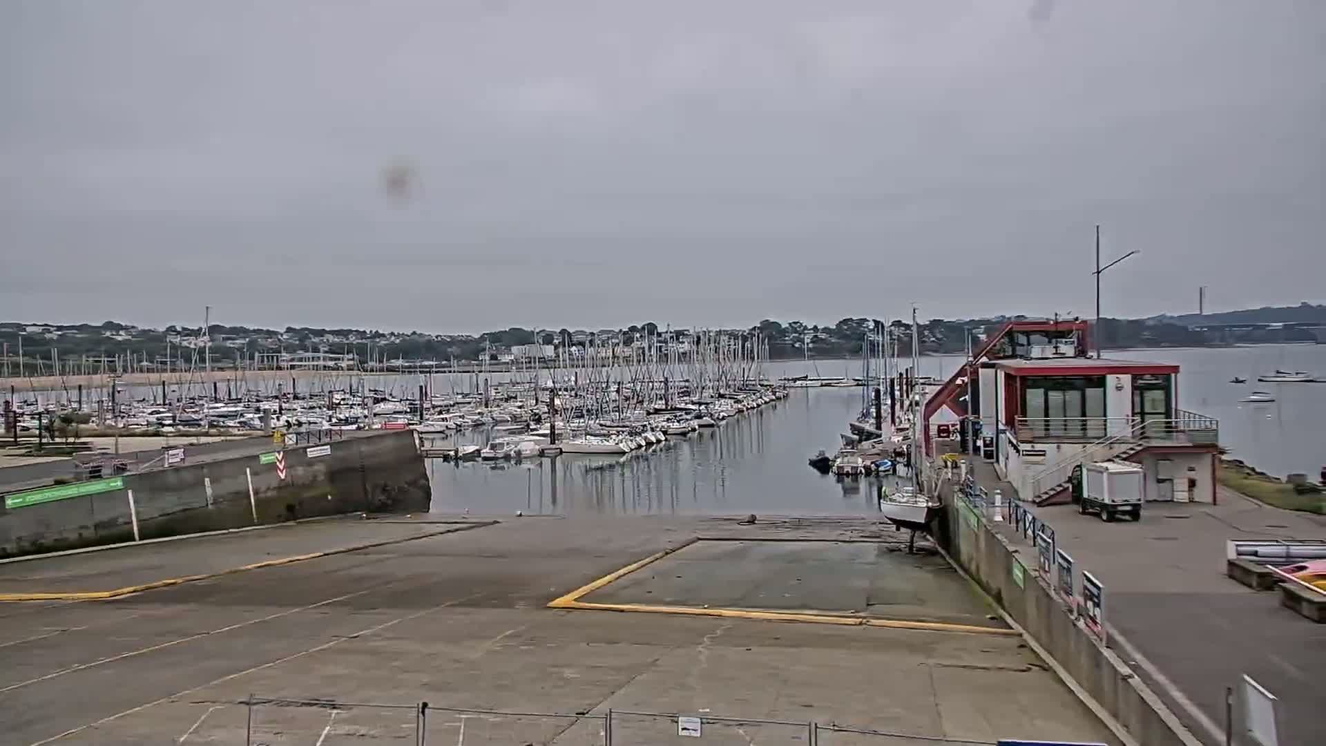 A marina filled with numerous sailboats is seen under an overcast sky, with a small building and paved area in the foreground.