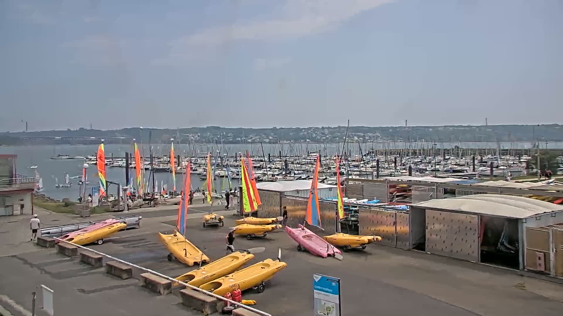 A marina filled with numerous sailboats and yachts under a partly cloudy sky, with several small sailboats and kayaks on land nearby.