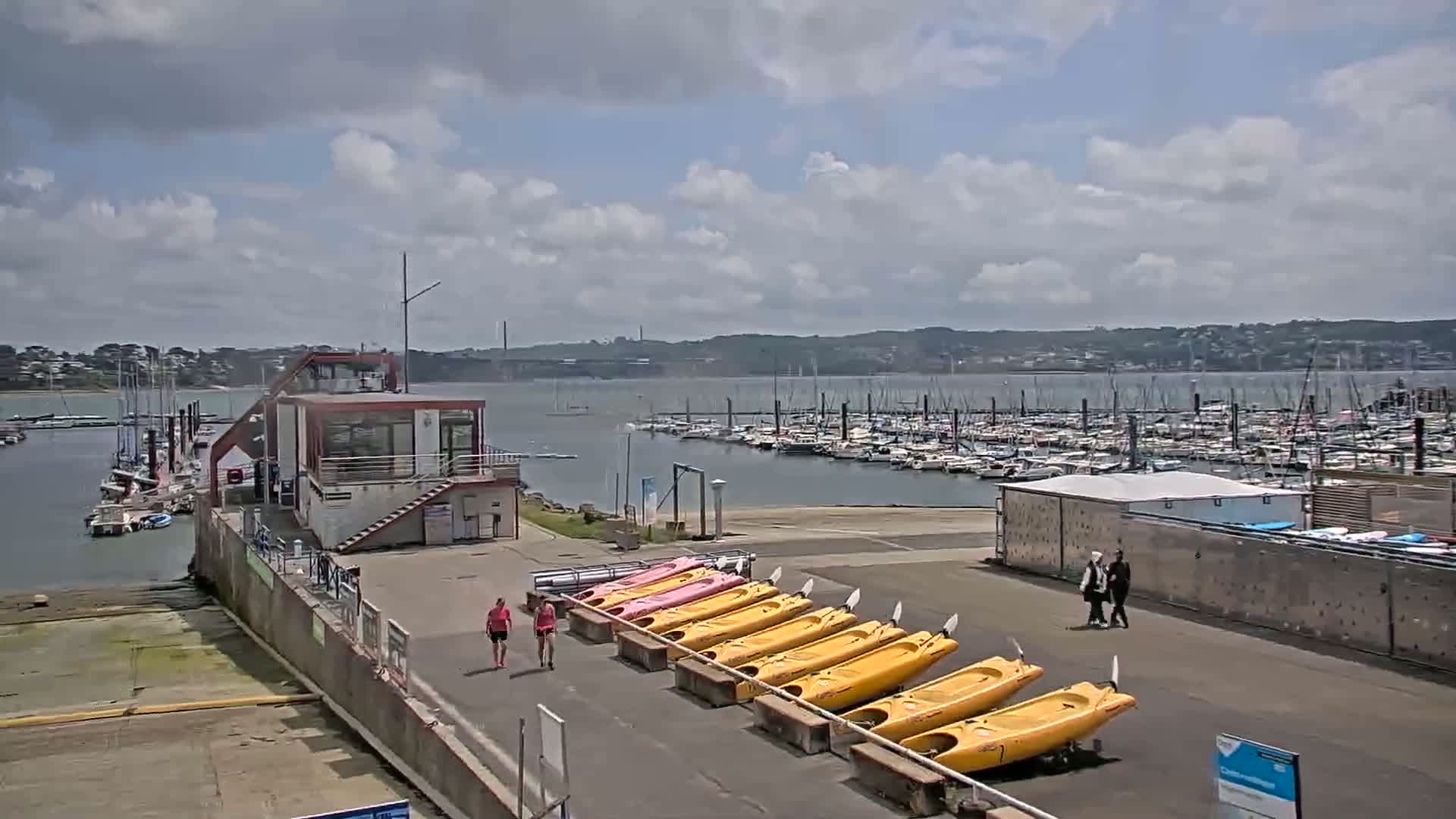 Under a partly cloudy sky, a marina with numerous boats is shown, with kayaks lined up on the shore near a small building and a few people walking by.