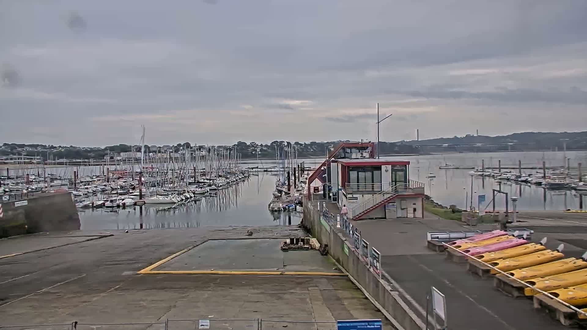A marina filled with sailboats under a cloudy sky, with a small building and kayaks visible in the foreground.
