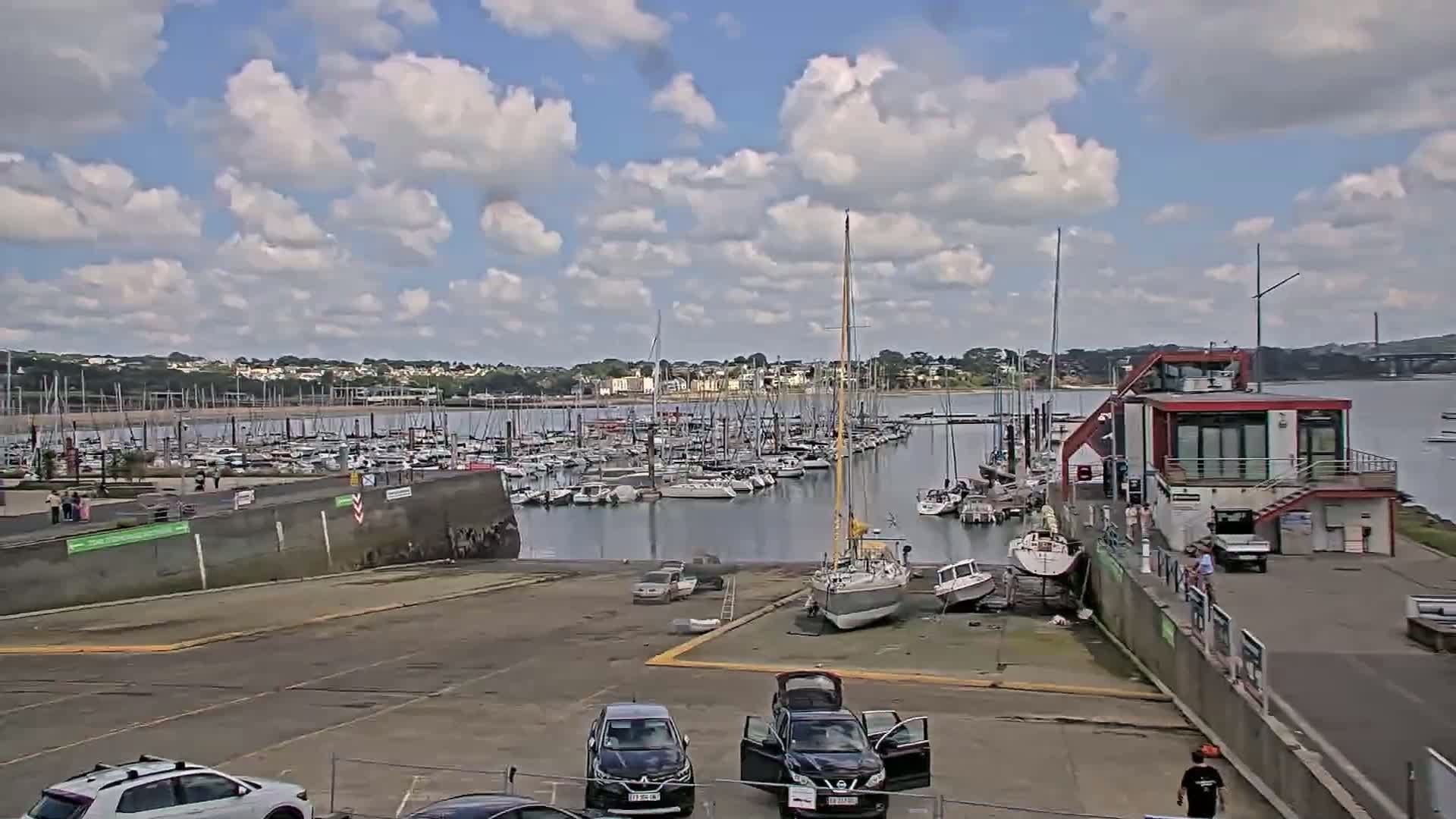 A marina filled with numerous sailboats and motorboats under a partly cloudy sky, with several cars parked in a lot nearby and a small building on the right.