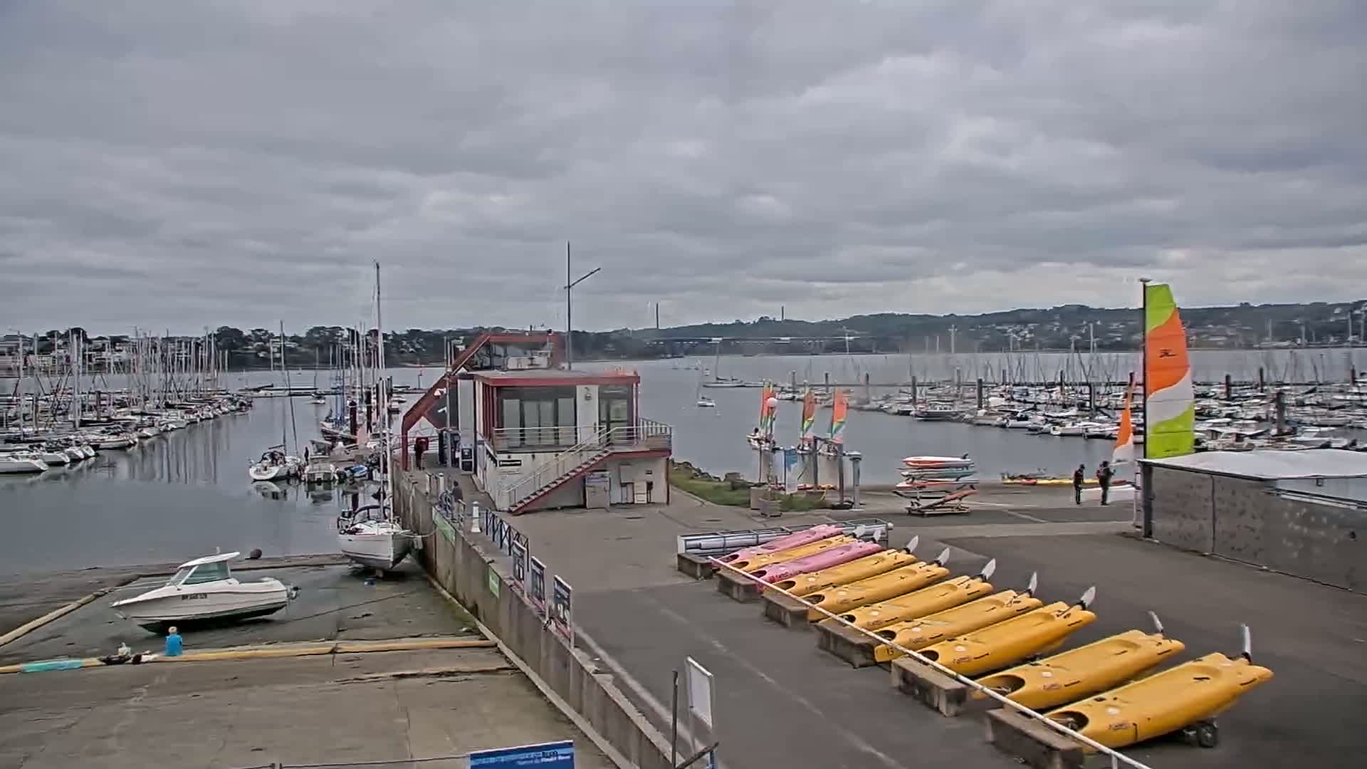 A marina filled with sailboats under a cloudy sky, with a small building, several kayaks, and a few people near the shore.