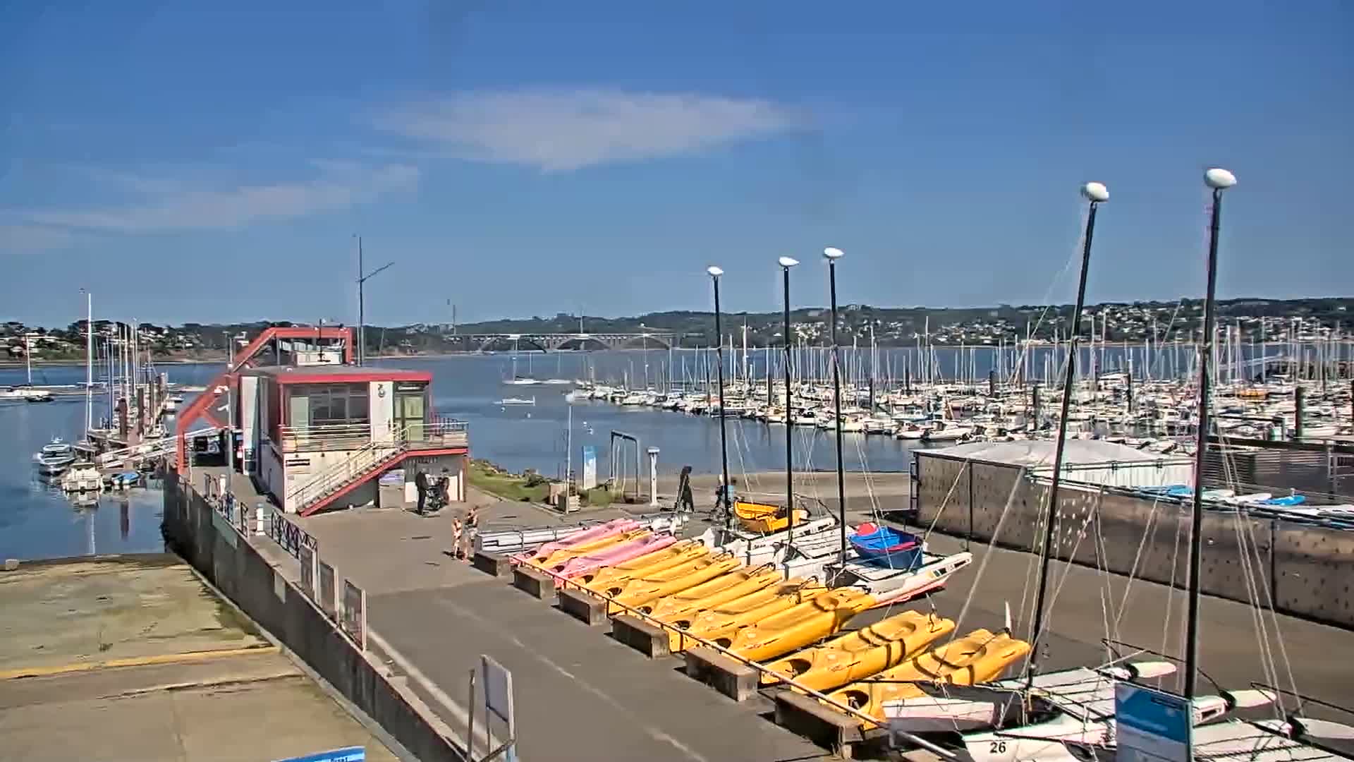 A marina with numerous sailboats and kayaks is shown on a sunny day, with a small building and a bridge visible in the background.