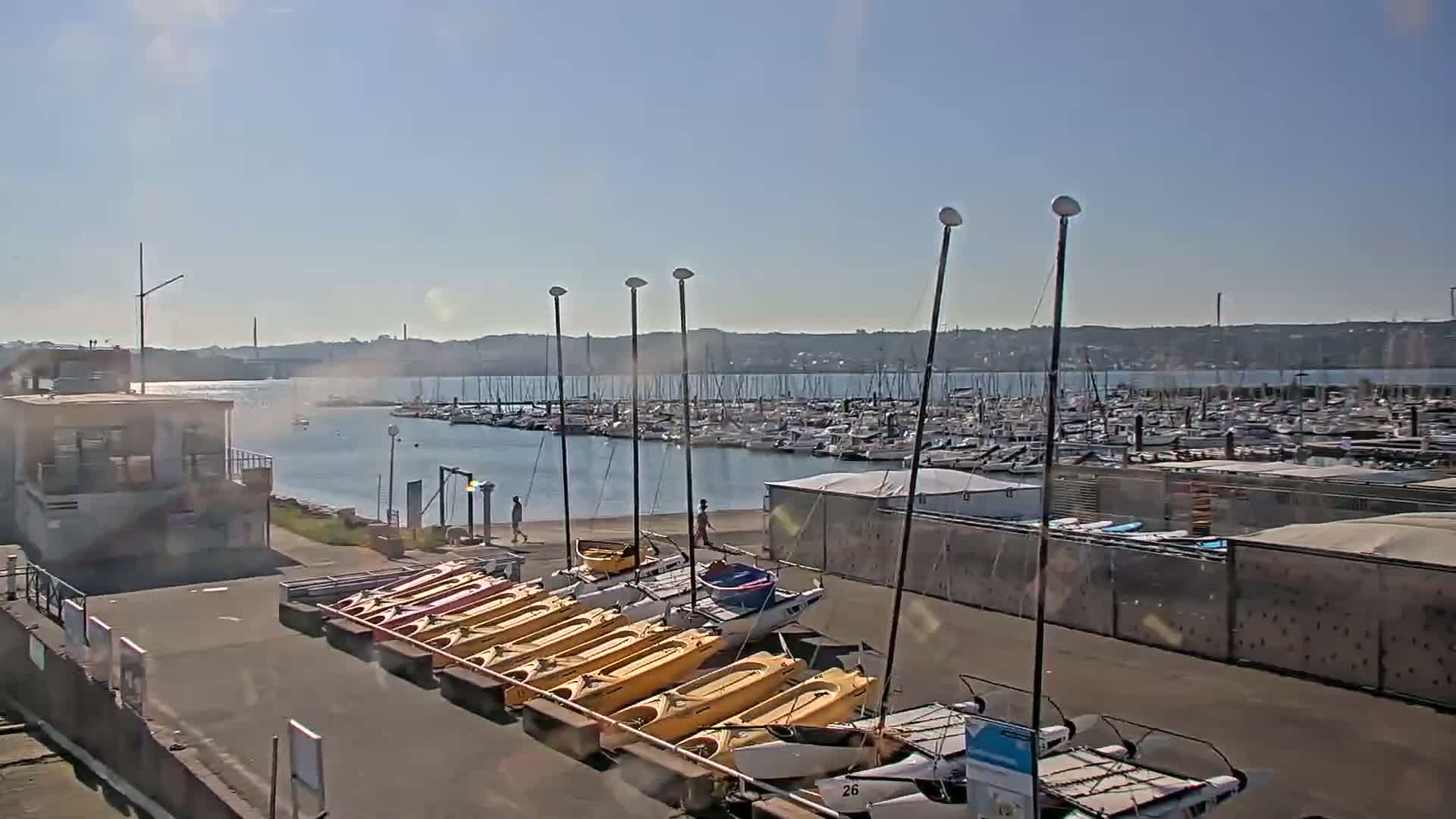 A marina on a sunny day, filled with numerous sailboats and small watercraft, including kayaks and catamarans, is bordered by a paved area and a small building.