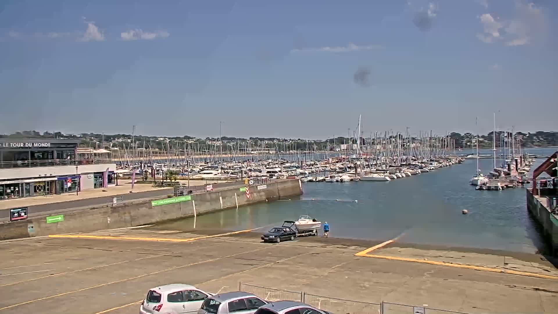 A marina filled with numerous sailboats and motorboats is seen on a sunny day, with a few cars parked in a lot nearby and a boat being launched into the water.