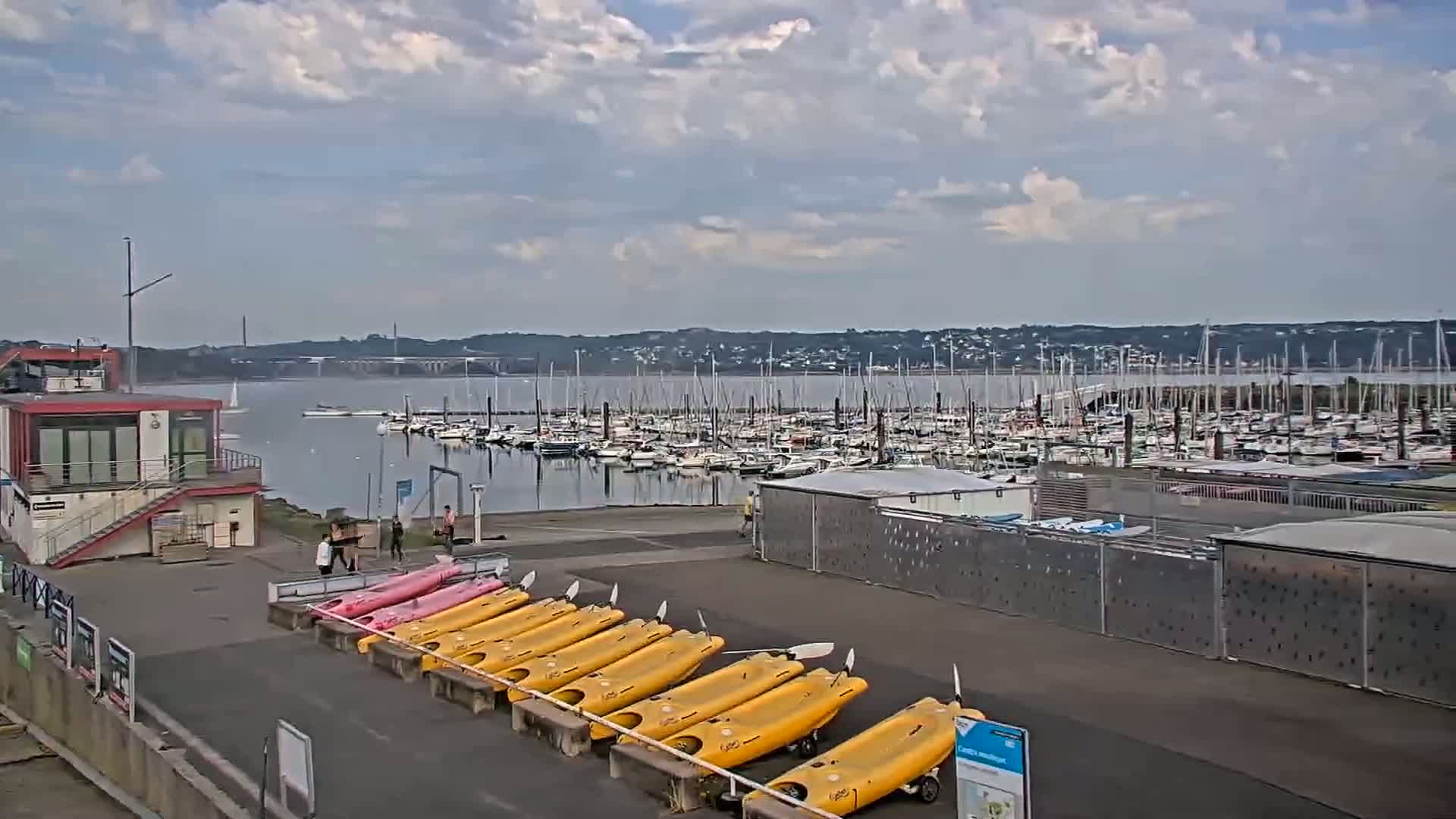 A marina filled with sailboats under a partly cloudy sky, with kayaks lined up on a nearby paved area.