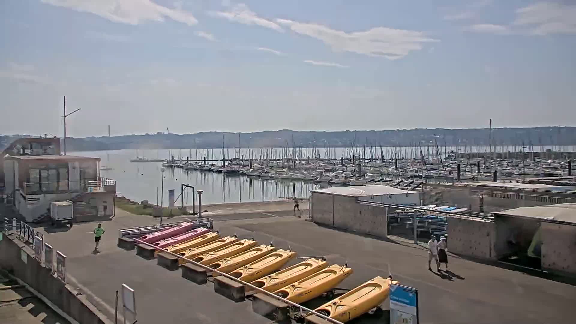 A marina filled with sailboats and a row of kayaks sits on a calm, sunny day near a building and several people.