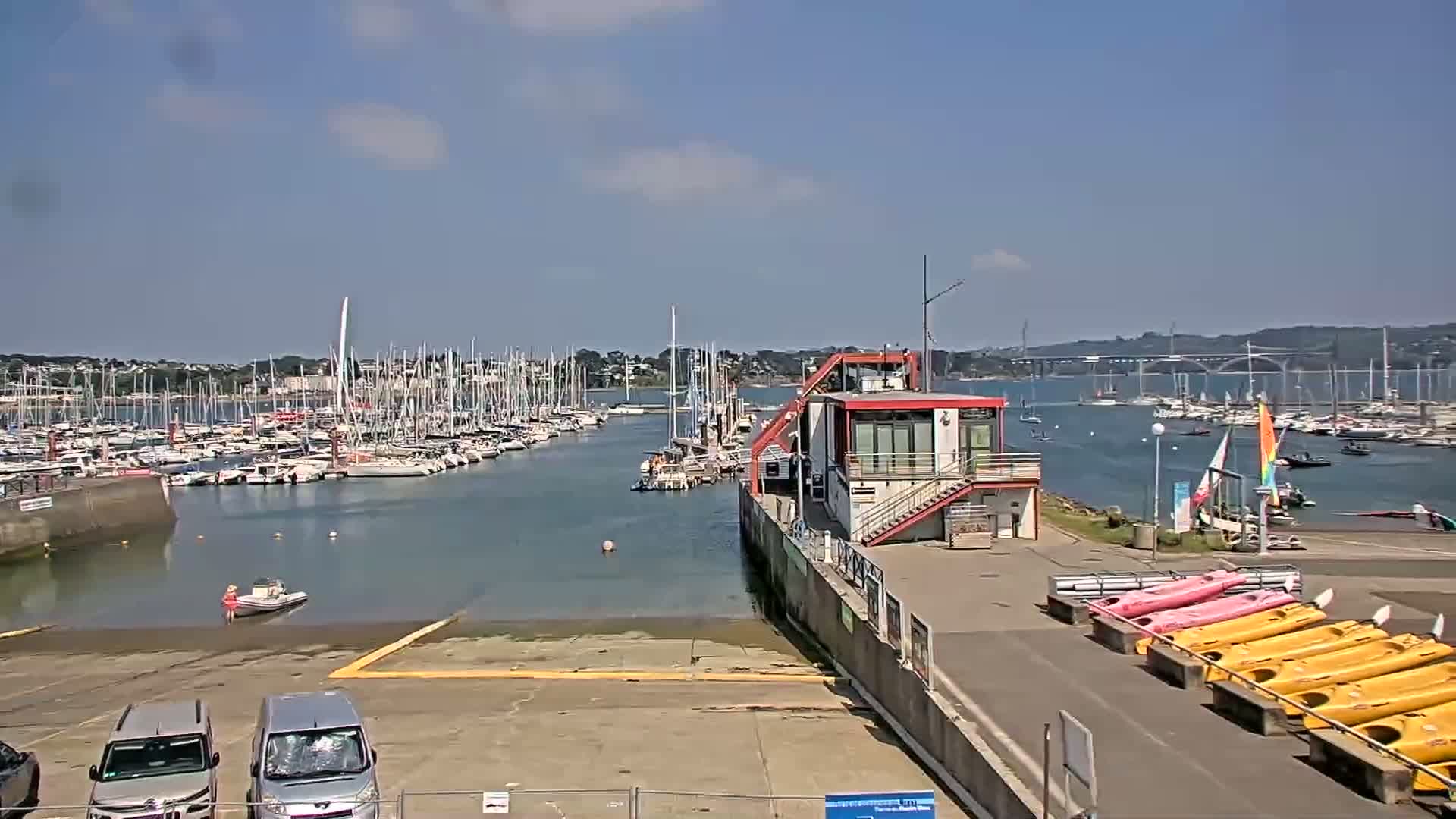 A marina filled with sailboats on a sunny day, with a small building and several kayaks visible near a boat ramp.