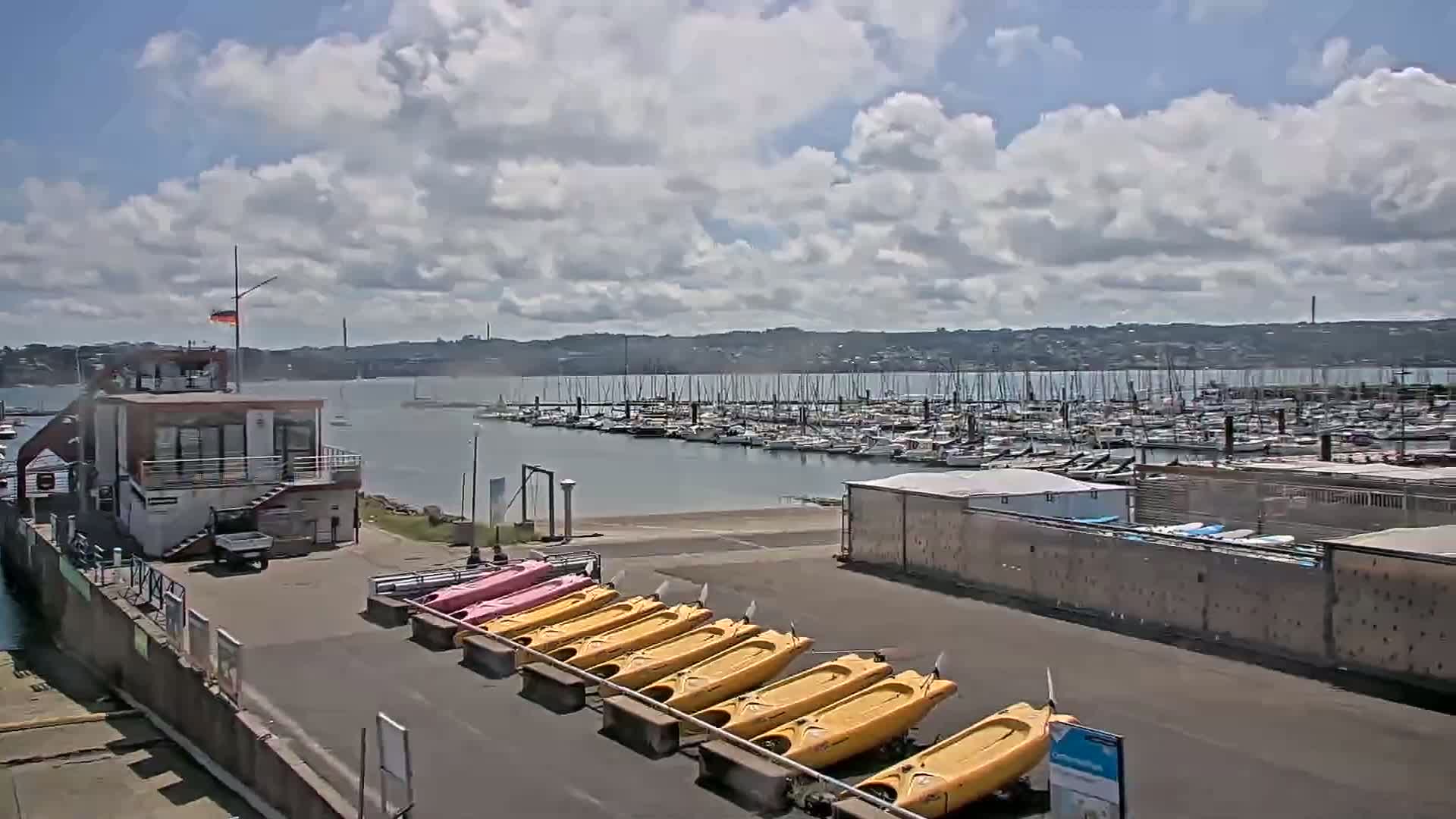 A marina with numerous sailboats and a row of kayaks sits under a partly cloudy sky.