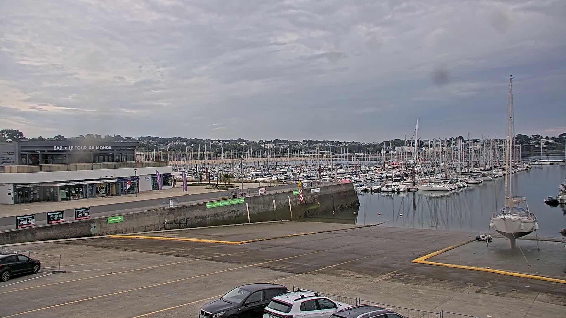 A marina filled with numerous sailboats under a cloudy sky, with a building and parking lot in the foreground.