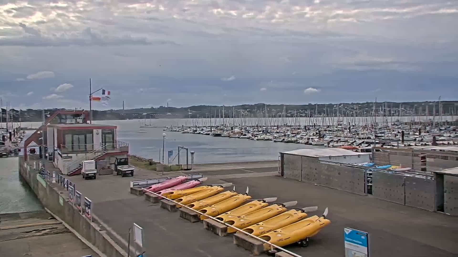 A marina with numerous sailboats and motorboats under a cloudy sky, featuring a small building and rows of kayaks on the shore.