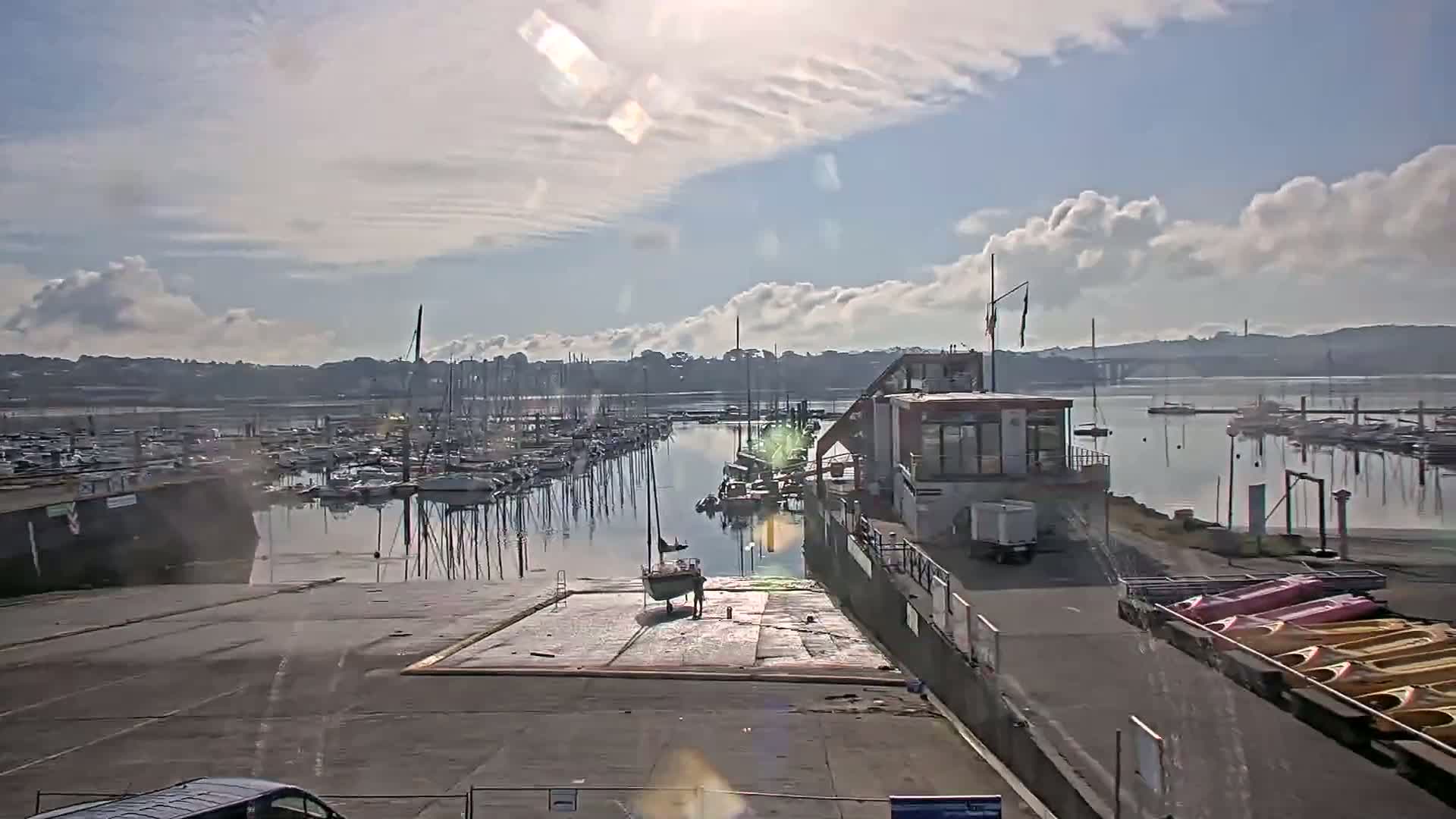 A marina filled with sailboats under a partly cloudy sky, with a small sailboat being pulled onto a slip and kayaks on a trailer nearby.