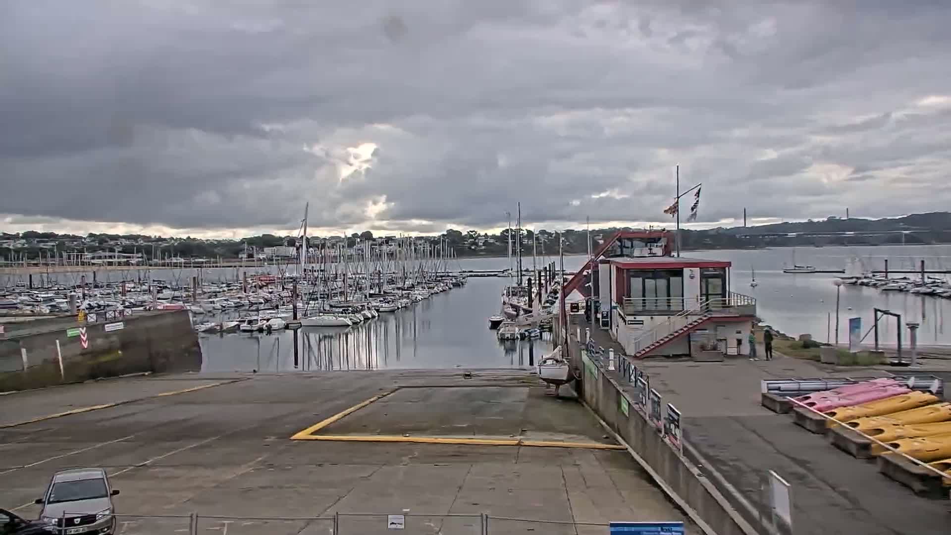 A marina filled with sailboats under a cloudy sky, with a boat ramp and a small building nearby.