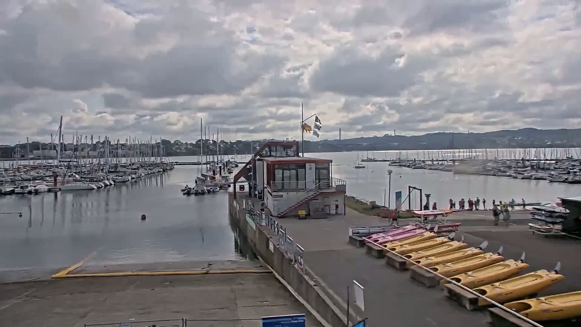 A marina filled with sailboats under a cloudy sky, with kayaks and a small building on the shore.