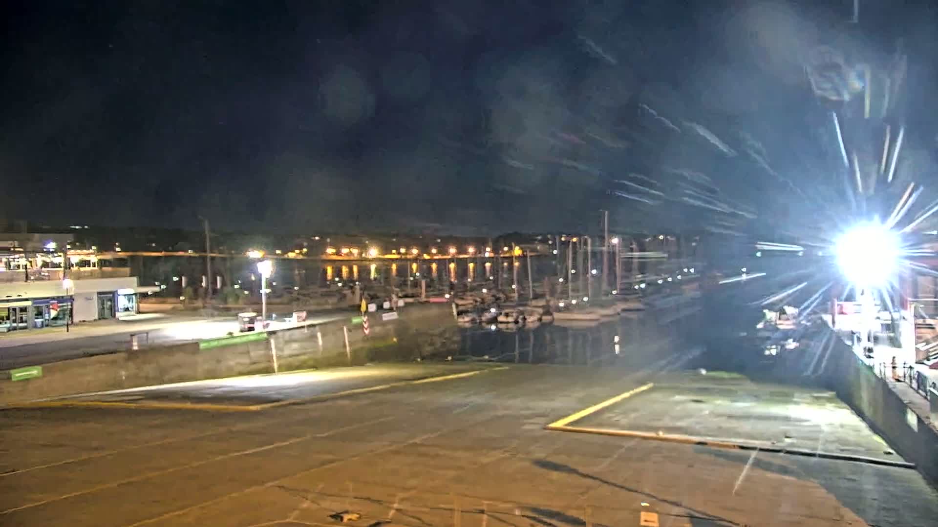 A nighttime view of a marina with docked sailboats under a dark, possibly slightly hazy sky, shows an empty boat launch area in the foreground.