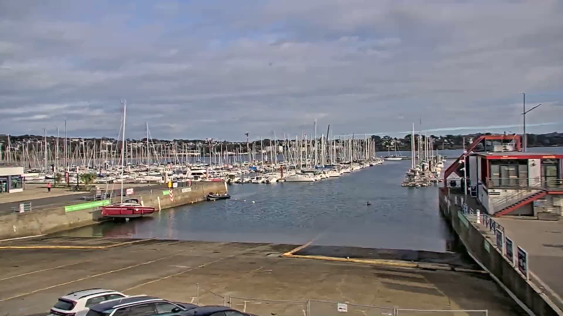A nighttime view of a marina with docked sailboats under a dark, possibly slightly hazy sky, shows an empty boat launch area in the foreground.