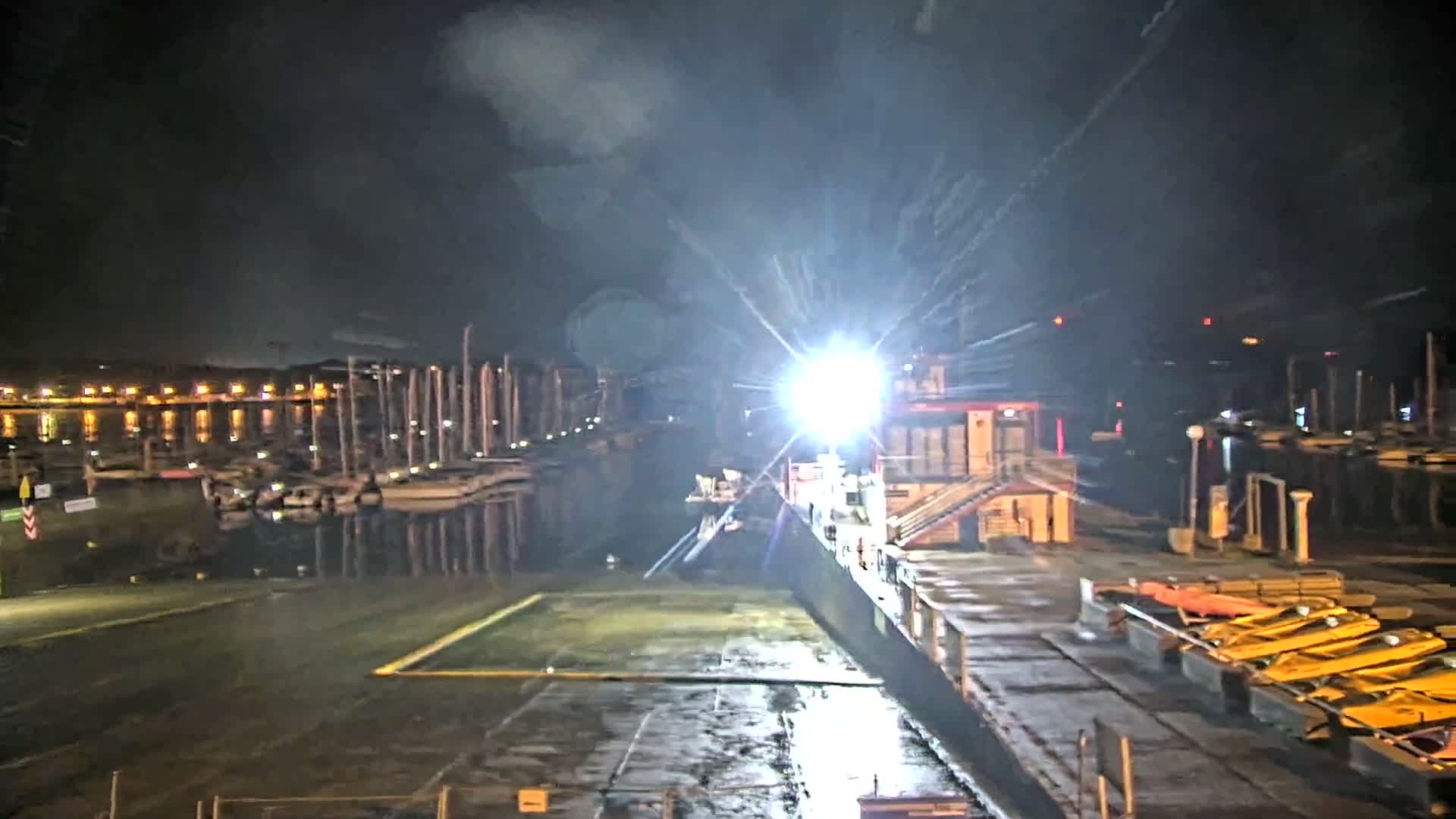 A powerful spotlight shines over a wet dock and marina filled with numerous boats under a dark, cloudy night sky.