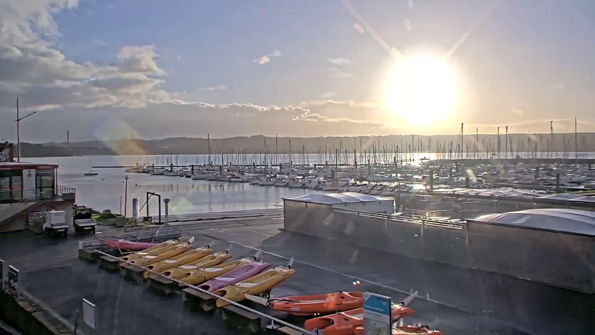 A sunny, partly cloudy day illuminates a bustling marina filled with numerous docked sailboats and a line of colorful kayaks on the shore, with distant land visible across the water.