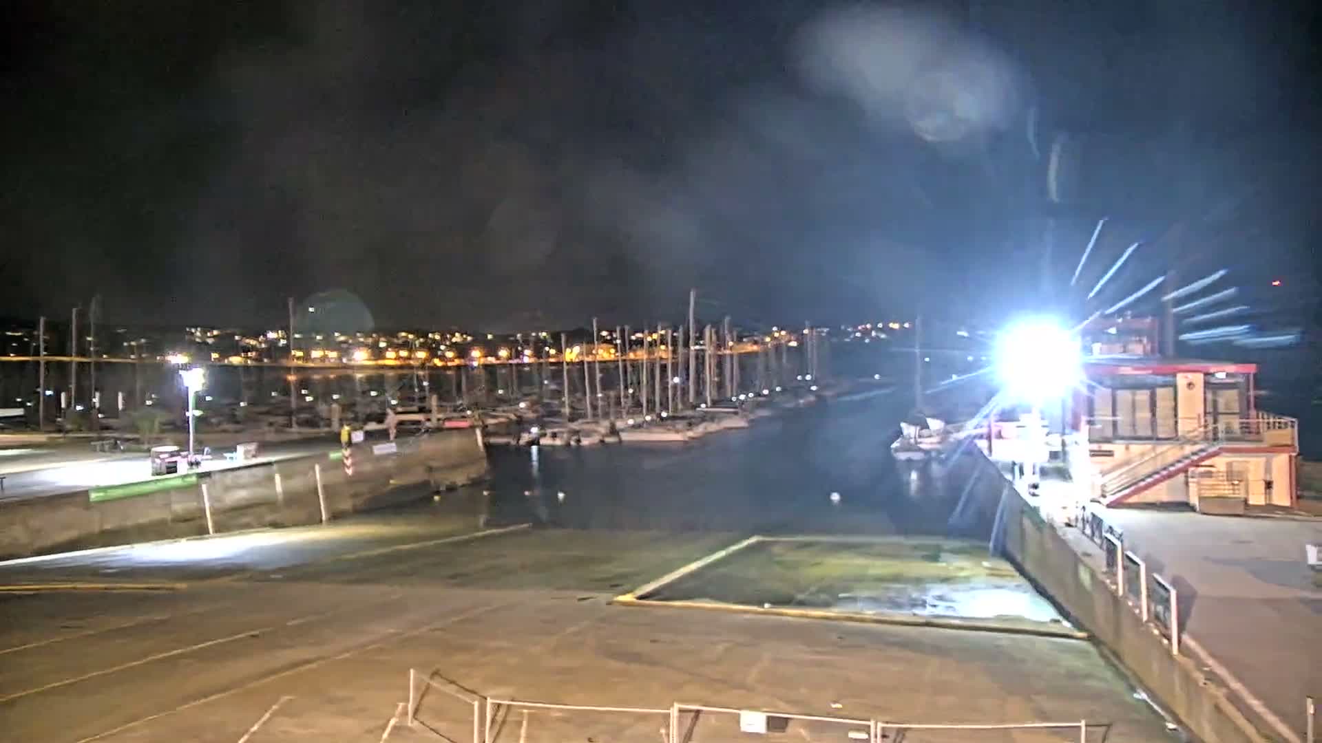 A sunny, partly cloudy day illuminates a bustling marina filled with numerous docked sailboats and a line of colorful kayaks on the shore, with distant land visible across the water.
