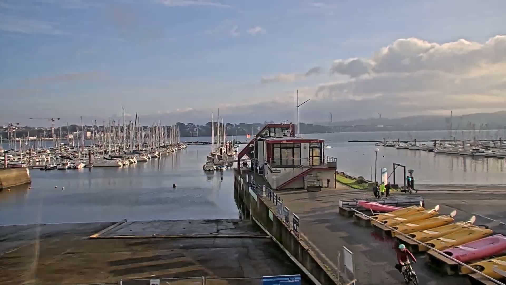 A sunny, partly cloudy day illuminates a bustling marina filled with numerous docked sailboats and a line of colorful kayaks on the shore, with distant land visible across the water.