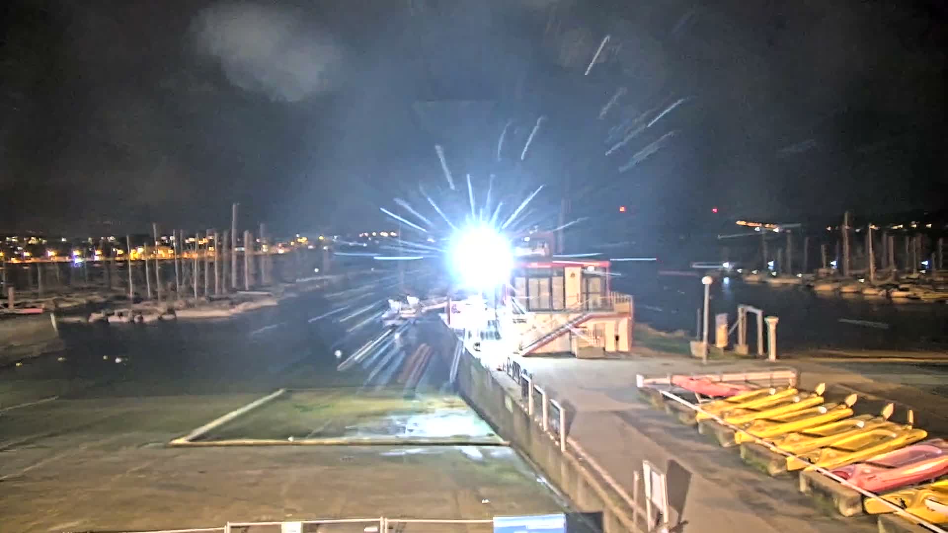 A sunny, partly cloudy day illuminates a bustling marina filled with numerous docked sailboats and a line of colorful kayaks on the shore, with distant land visible across the water.