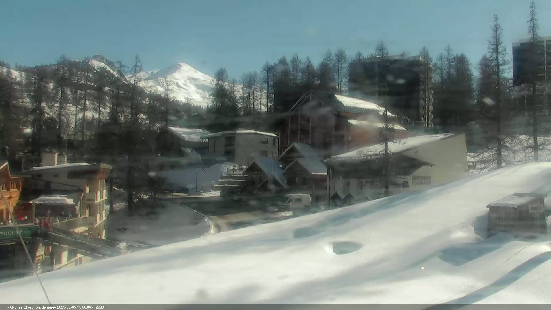 A snow-covered village nestled among snow-dusted evergreen trees, with a bright, sunny sky and mountains in the background.