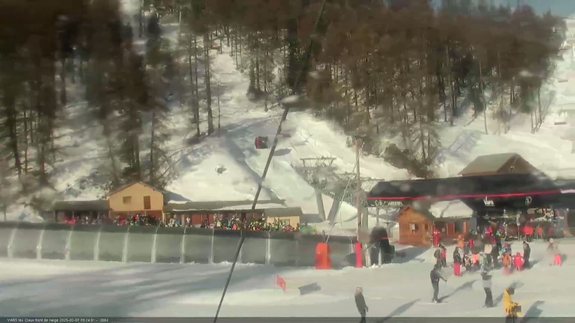 A sunny day at a snow-covered ski resort shows a ski lift, several buildings, and many people gathered near a snow-covered slope.