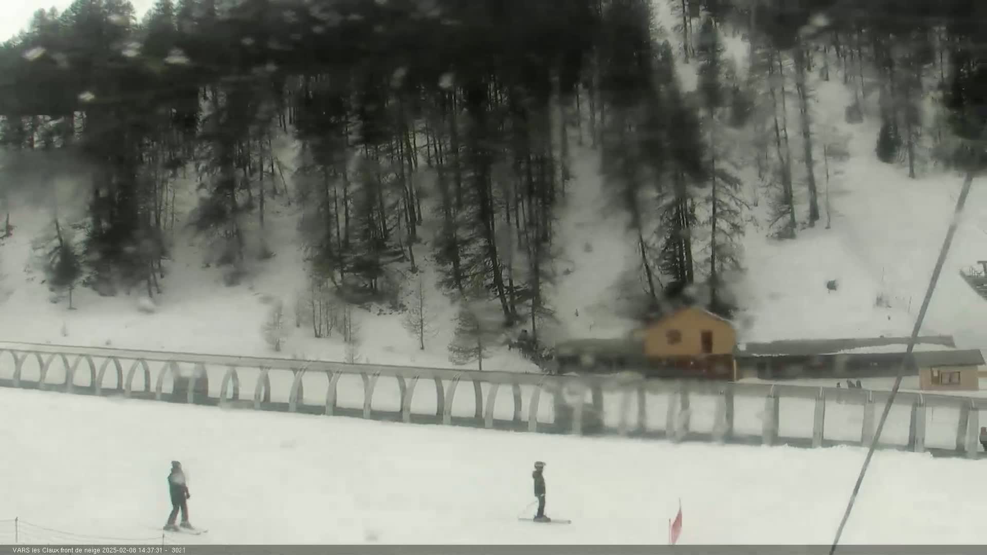 Two skiers are on a snowy slope below a snow-covered forest and a building, with a fence separating the slope from the woods.