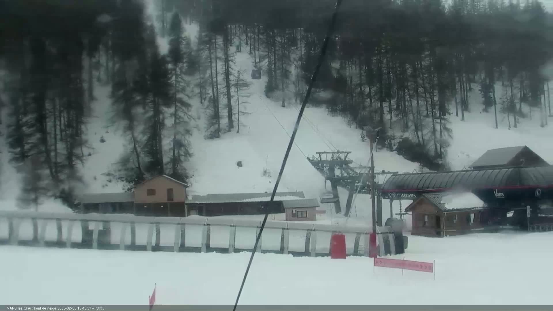 A snow-covered ski slope with a chairlift, several small buildings, and a line of trees in the background during a snowstorm.