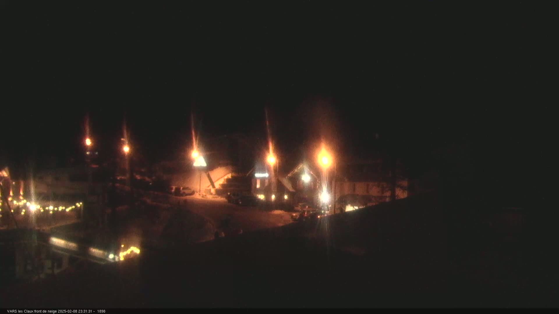 A nighttime snow-covered scene shows a building complex illuminated by streetlights and string lights.