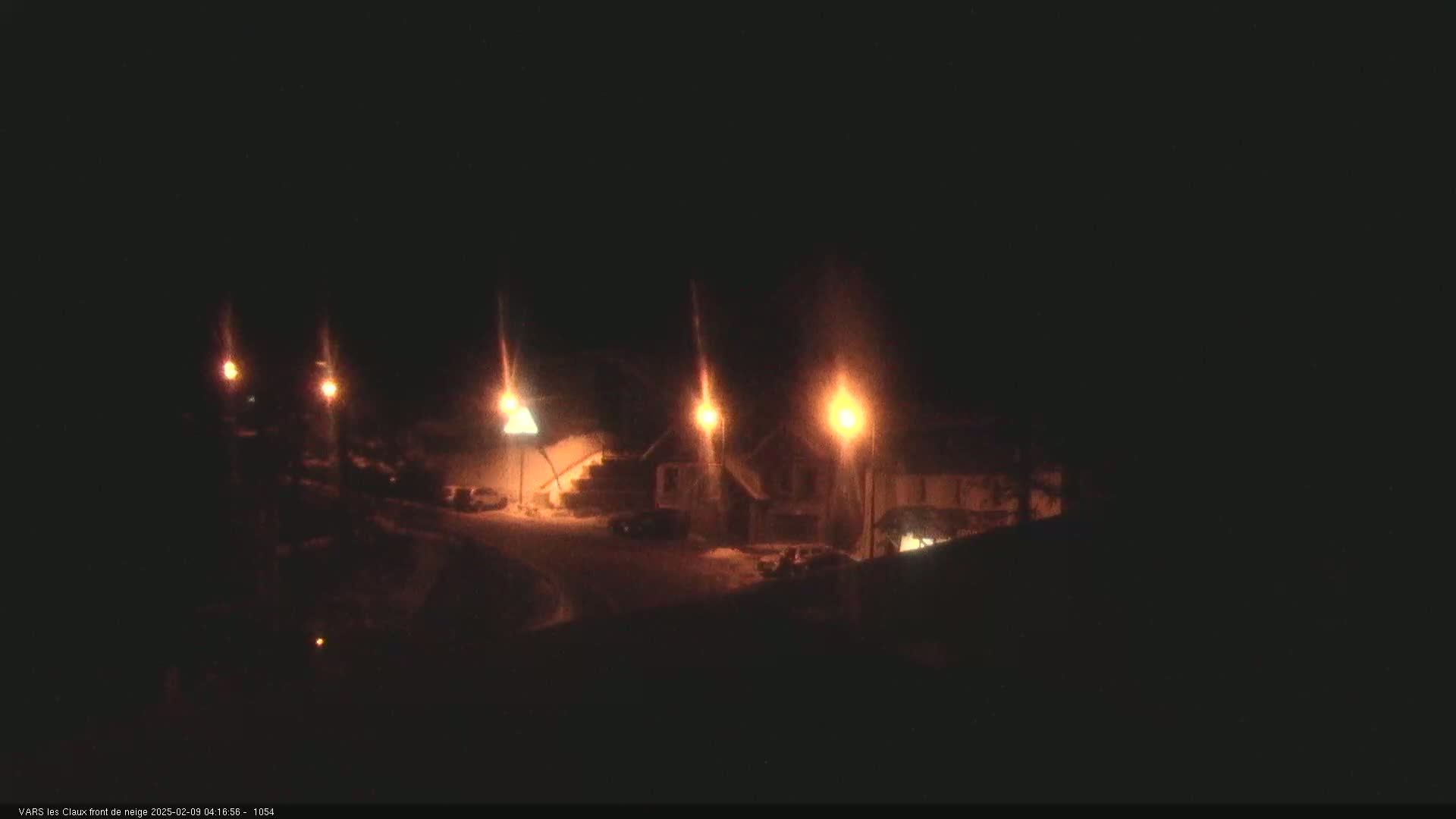 A nighttime scene of a snow-covered area with several buildings illuminated by streetlights.