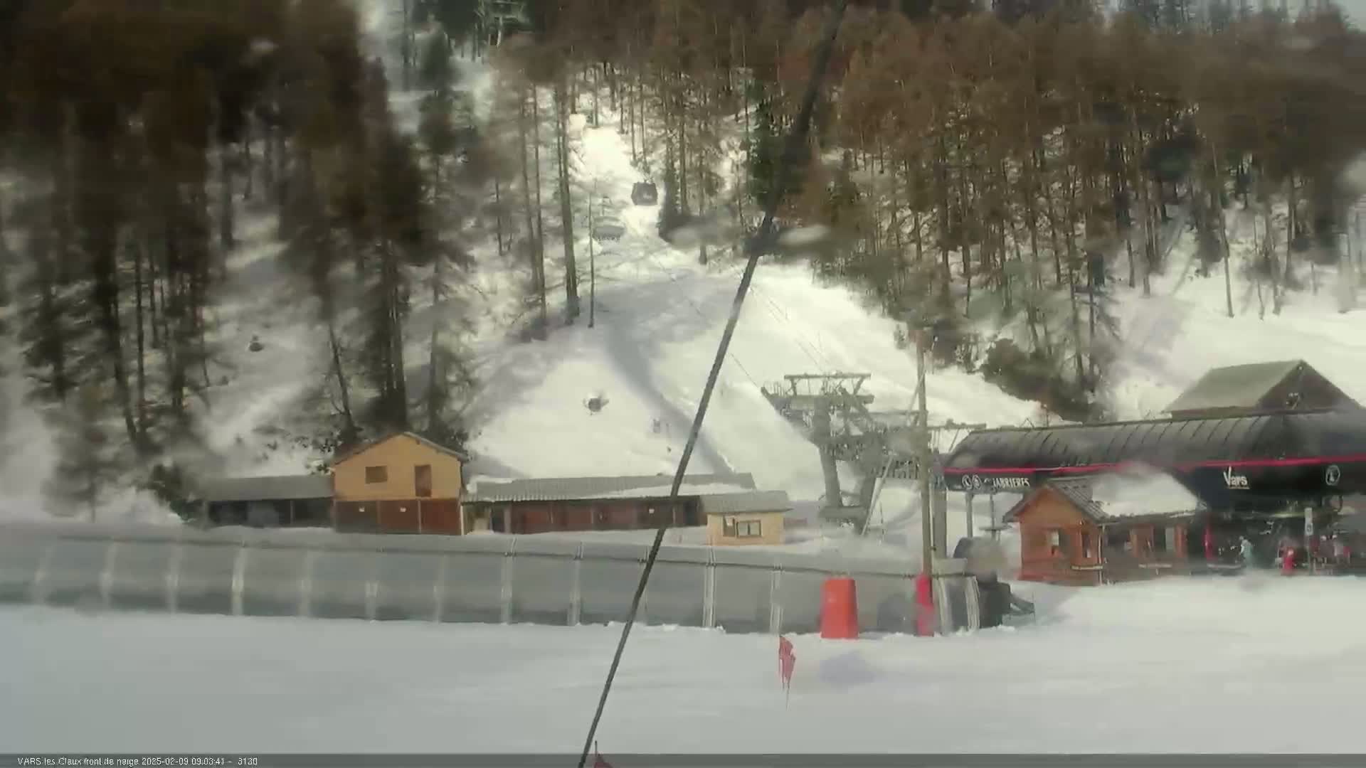 A snowy ski resort scene shows a chairlift, several small buildings, and a snowmaking machine operating amidst a coniferous forest under overcast conditions.
