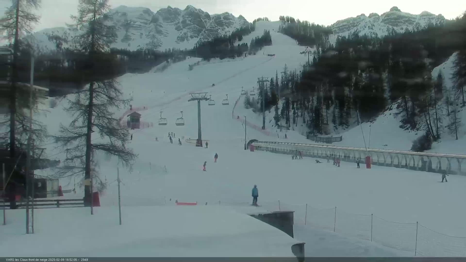 A snow-covered ski slope with several skiers and a chairlift is seen against a backdrop of snow-capped mountains under an overcast sky.