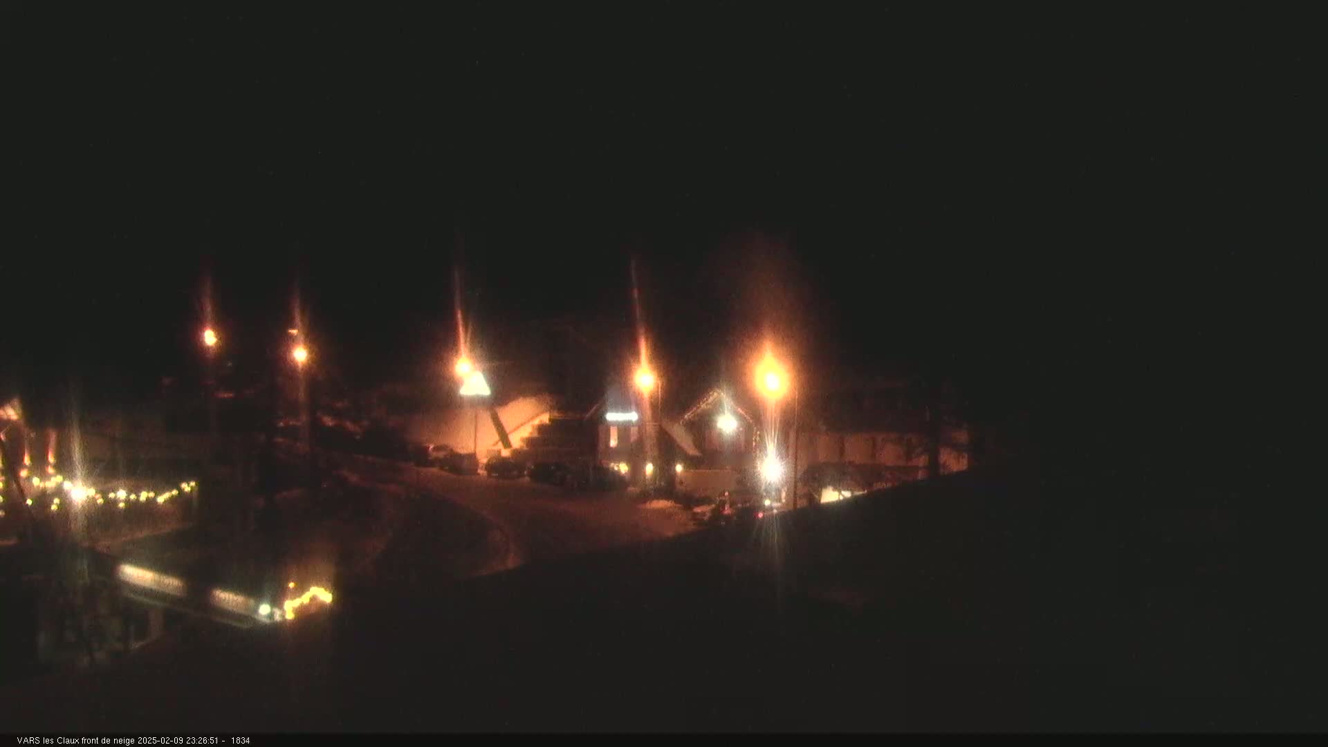 A nighttime view of a snow-covered area with illuminated buildings and string lights.