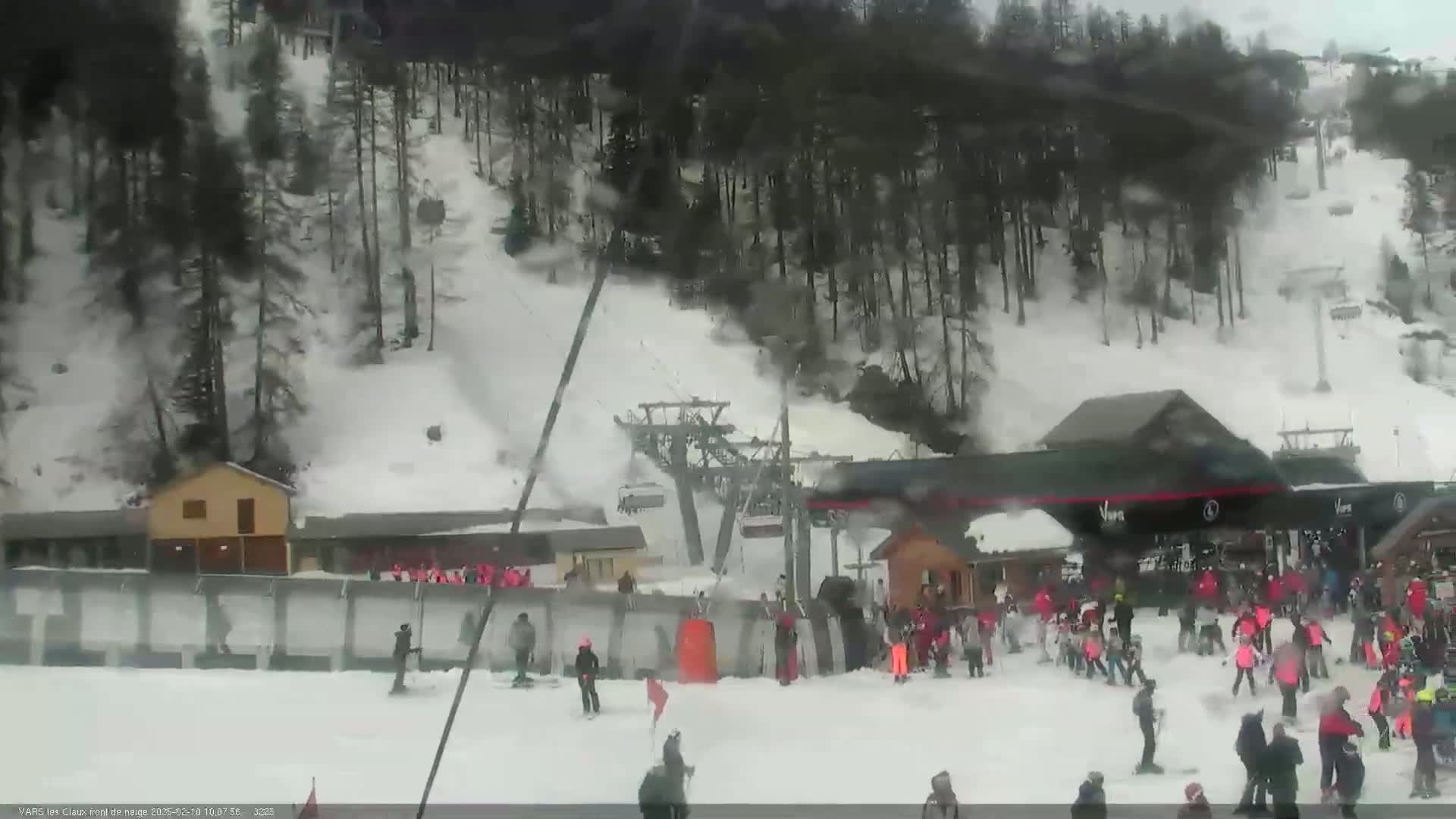 A snowy ski resort with a chairlift and many skiers and snowboarders at the base, under a cloudy sky.