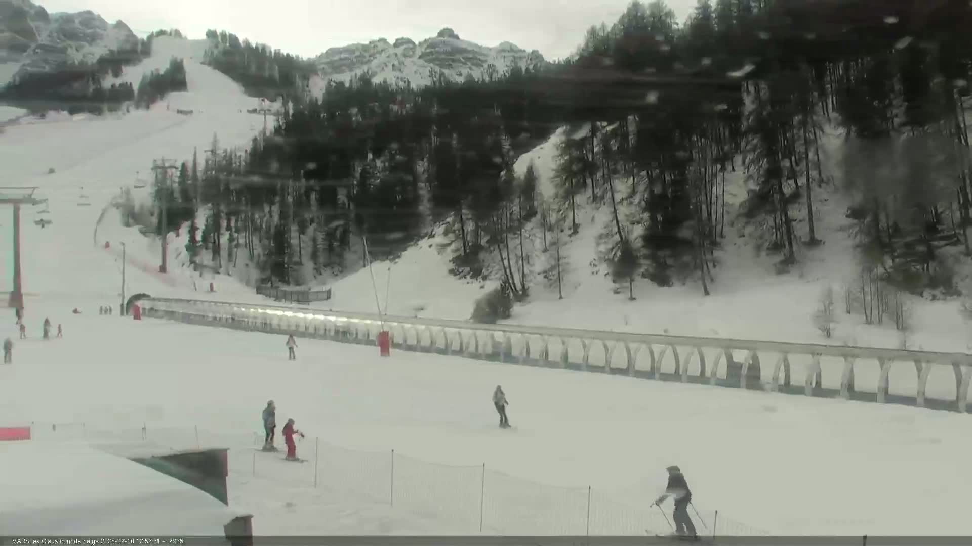 A snowy ski slope with several skiers, a long lift, and a snow-covered forest in the background under an overcast sky.