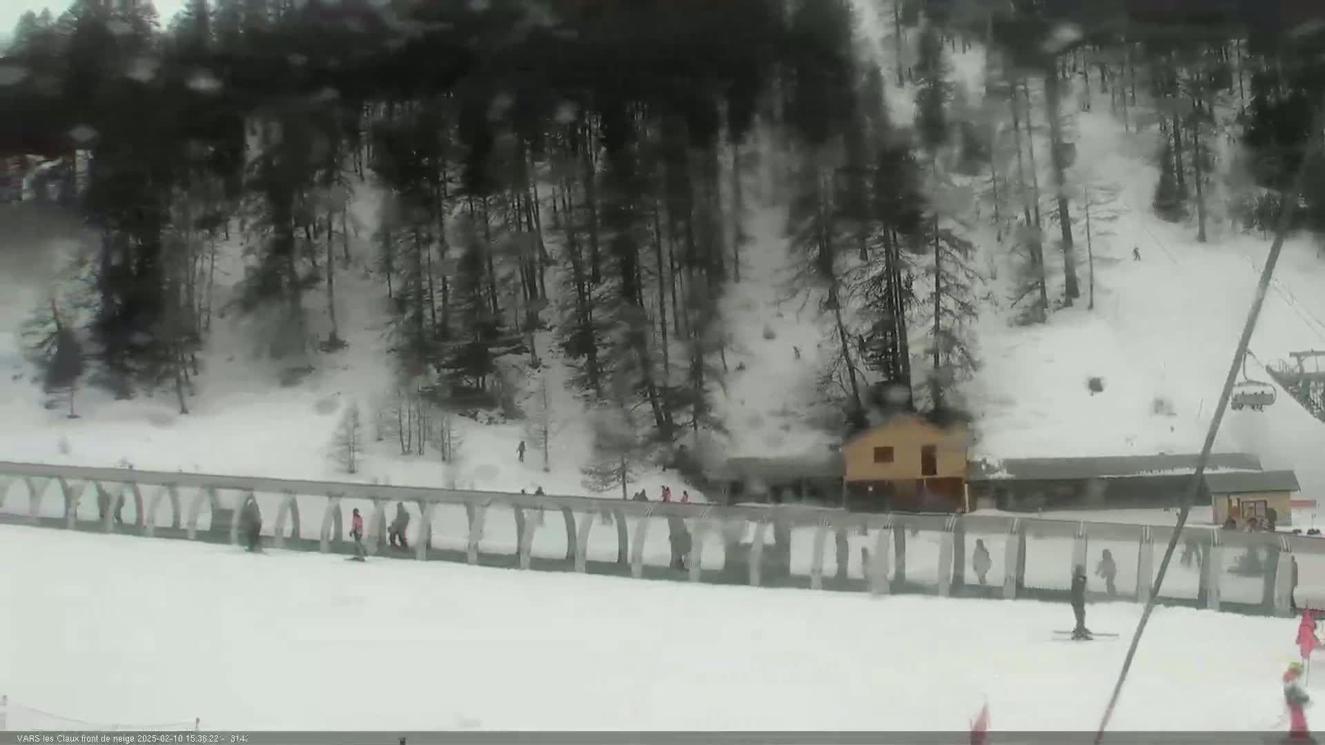 A snowy, overcast scene shows a ski slope with several skiers and snowboarders near a building nestled amongst a dark evergreen forest.