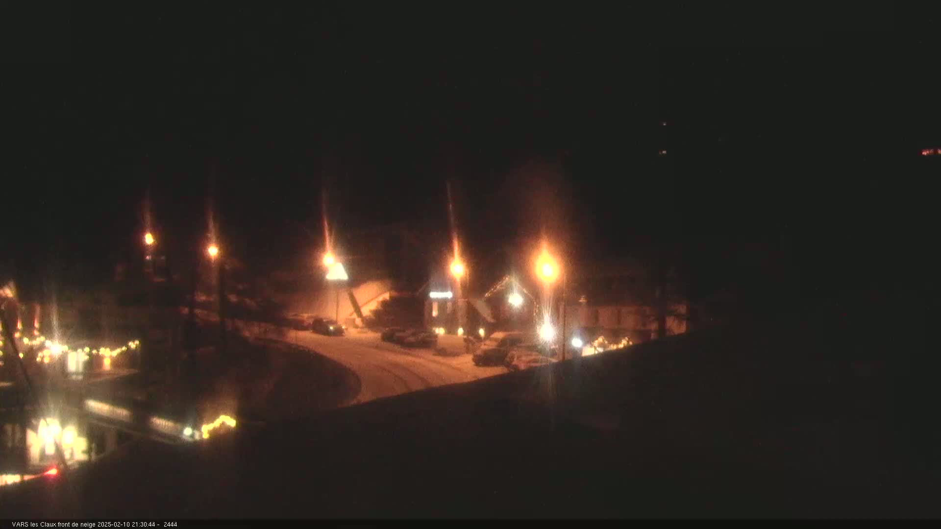 A nighttime, snow-covered street scene shows several buildings illuminated by streetlights and holiday lights, with cars parked along the street.