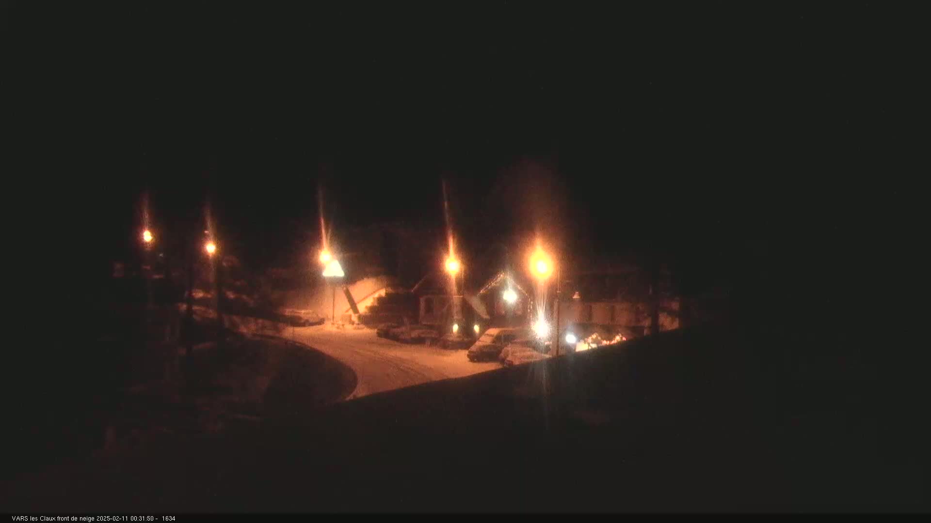 A snow-covered parking lot with several buildings and cars is illuminated by streetlights at night.