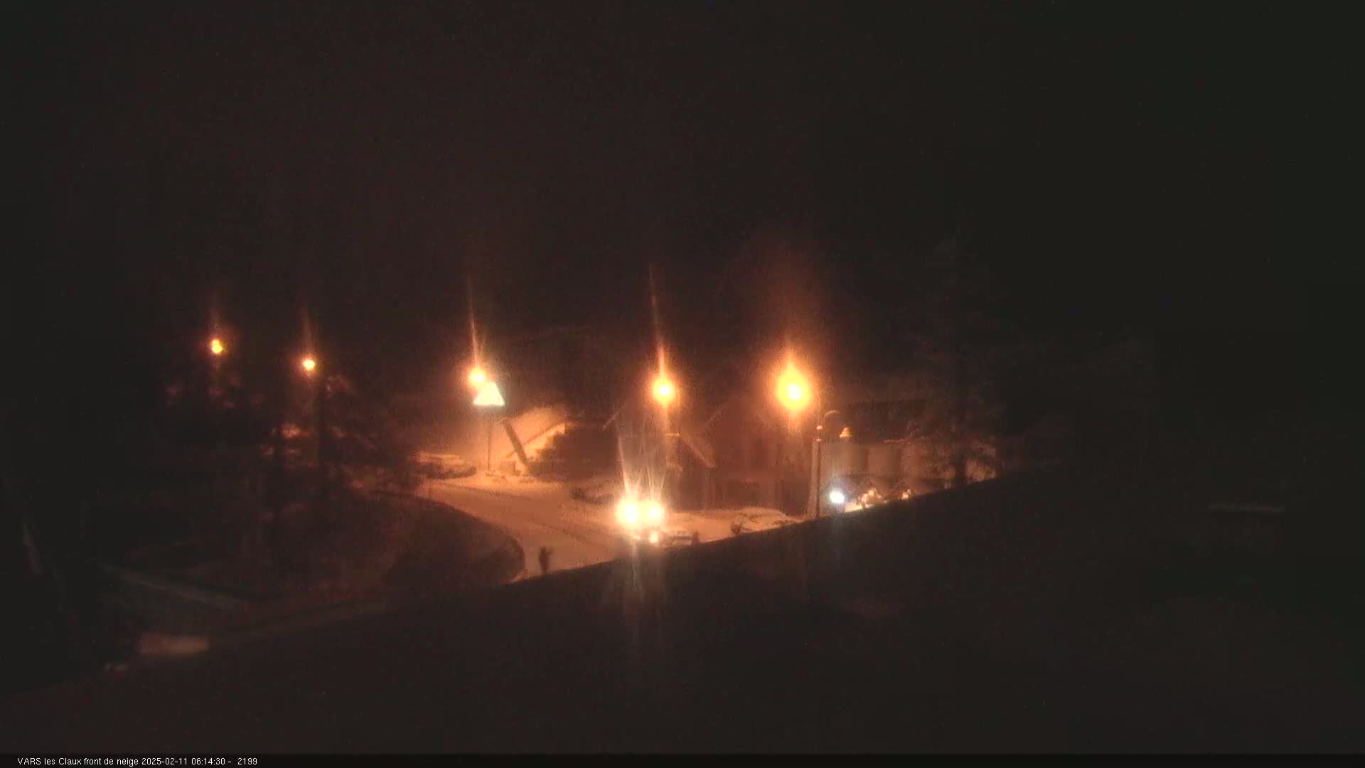 A nighttime, snowy scene shows a road with illuminated streetlights and a few vehicles near buildings.