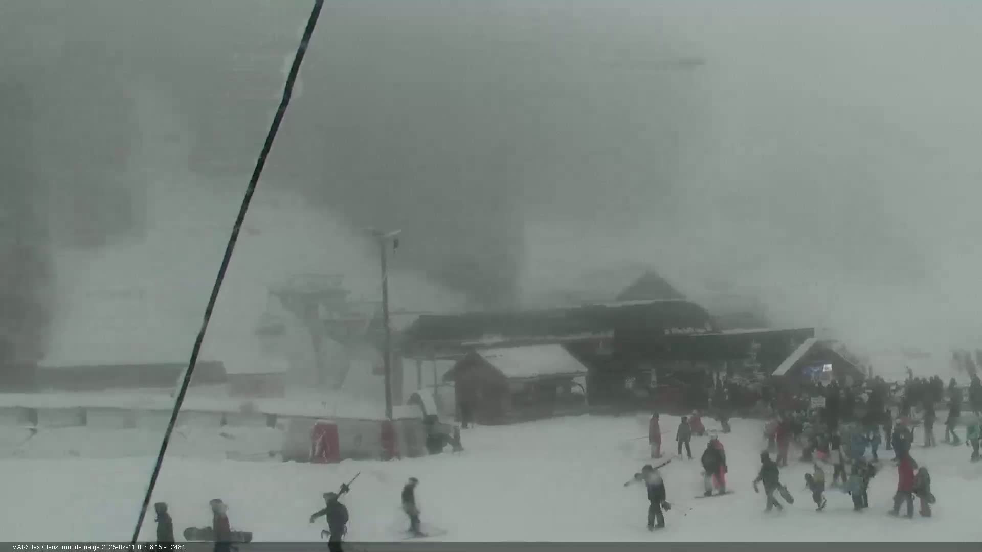 A snowy scene at a ski resort shows a group of people bundled in winter clothing, with a building and ski lift visible through heavy snowfall and blowing snow.