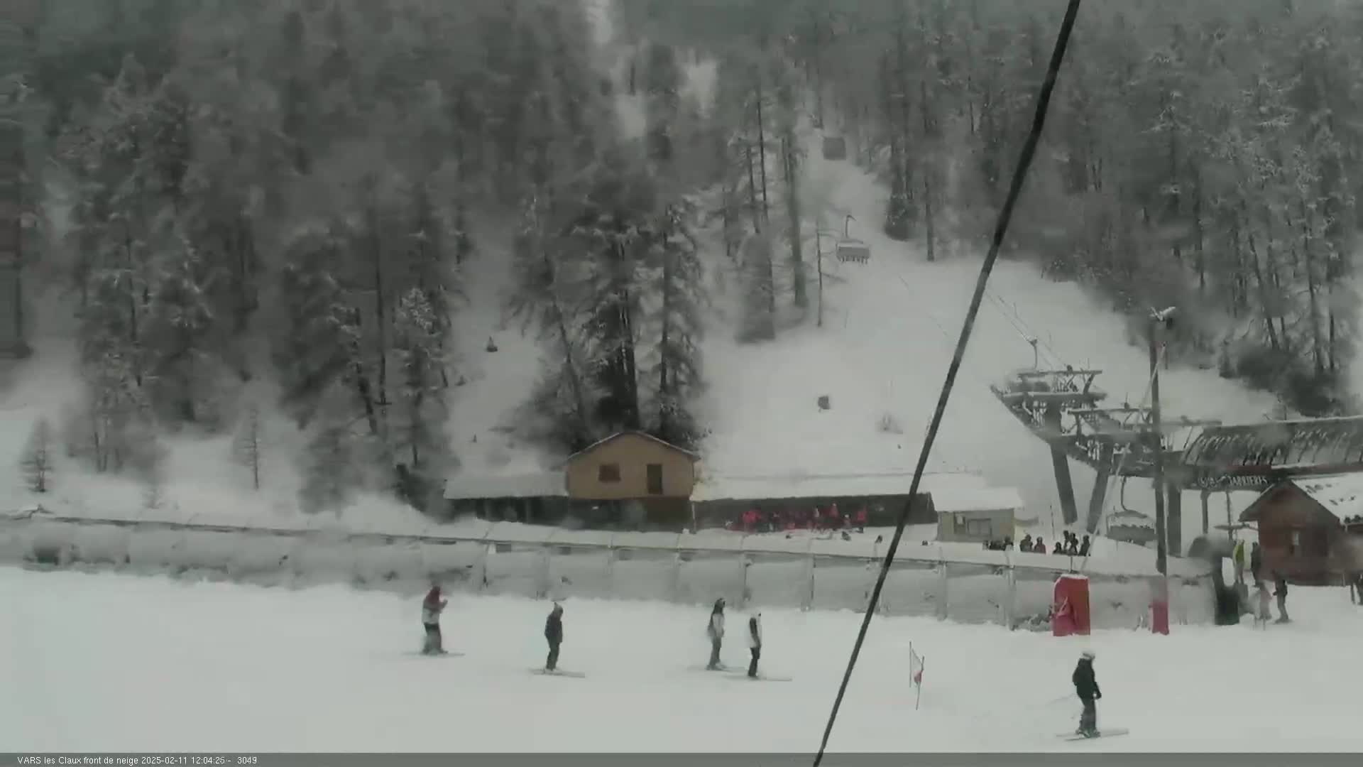 A snowy ski slope with a chairlift and several skiers, amidst snow-covered trees and buildings in a snowstorm.