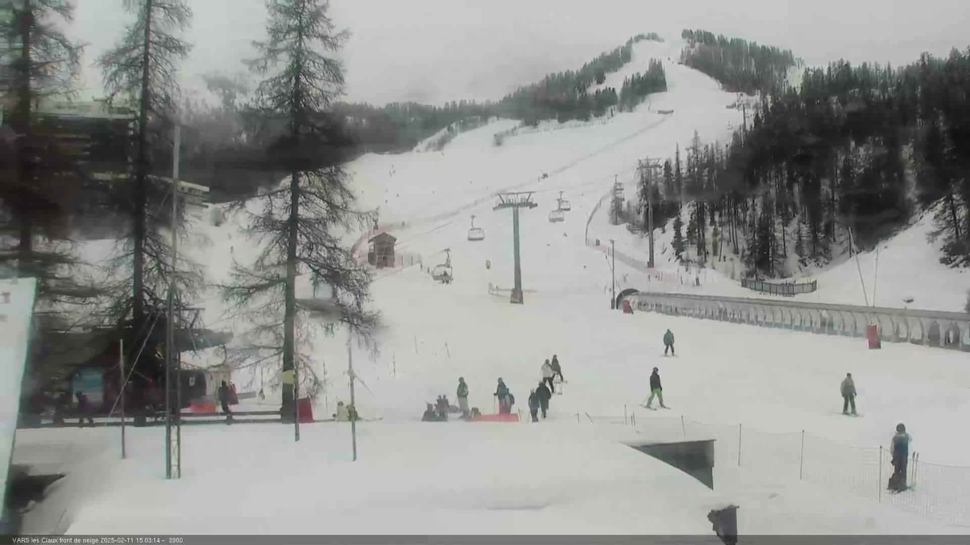 A snowy ski slope with a chairlift and several skiers visible under an overcast sky.
