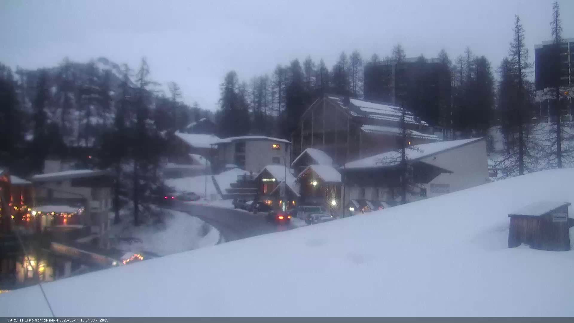 A snow-covered village scene at dusk shows several buildings and vehicles near a curving road, with evergreen trees in the background.